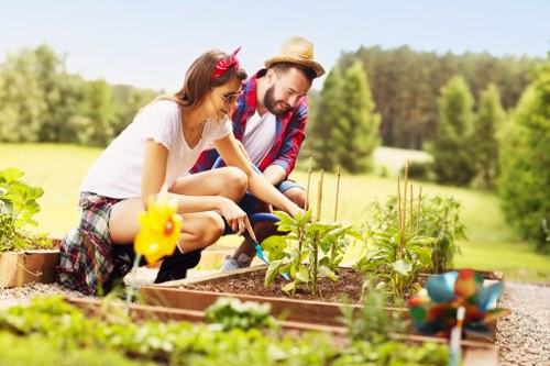 The image depicts a couple engaged in gardening within a well-maintained outdoor space, possibly in the garden of a property in St Johns Wood or nearby. The foreground features a raised garden bed filled with dark, rich soil, containing young green plants and small seedlings. The couple, a woman and a man, are actively tending to the plants, with the woman using garden tools and the man observing closely. They are dressed casually, suitable for outdoor gardening tasks, with the woman wearing a striped skirt and a white top, and the man in a plaid shirt and a straw hat. The background showcases a lush, expansive lawn with neatly cut grass, bordered by a row of mature trees and hedges, creating a natural and private outdoor environment. The scene is lit by natural daylight, indicating clear weather, and the overall setting emphasizes a tidy, landscaped garden ideal for outdoor leisure and gardening services by Gardening Services St Johns Wood. The image captures a moment of engagement with nature, highlighting typical garden features such as soil beds, greenery, and outdoor tools, with a focus on creating a nurturing and vibrant garden atmosphere in a residential outdoor space in London.