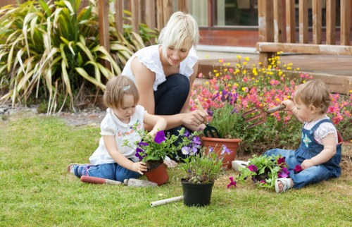 A woman and two young children are engaged in gardening activities in a well-maintained backyard garden, which features a lush green lawn, potted plants with colorful flowers, and a variety of flowering shrubs and plants. The scene is set on a bright, clear day with natural outdoor lighting. The woman is sitting on the grass, carefully tending to the potted plants, while the two children sit nearby, each holding small pots with blooming flowers, actively participating in the gardening process. In the background, there is a wooden deck with steps leading to the garden, framed by tall ornamental grasses and a garden fence. The overall environment illustrates a neat, landscaped outdoor space suitable for family gardening, supporting professional gardening and landscaping services offered by Gardening Services St Johns Wood in the NW8 postcode area, with natural tones of green, purple, pink, and yellow enhancing the gardenâ€™s inviting appearance.