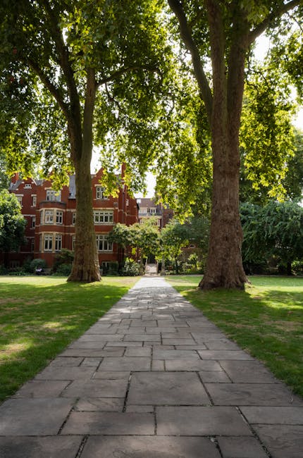 A paved stone pathway runs through a lush, well-maintained garden flanked by two large, mature trees with thick trunks and dense green foliage. The grass on either side of the path is neatly trimmed, 